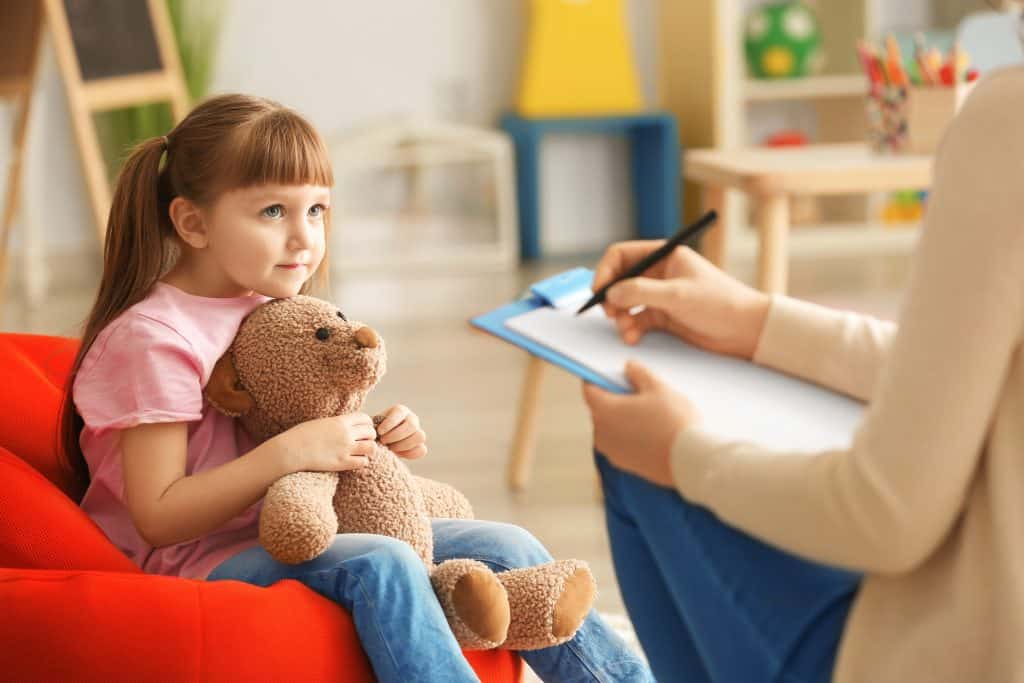 Niña con oso de peluche frente a una psicóloga que toma notas